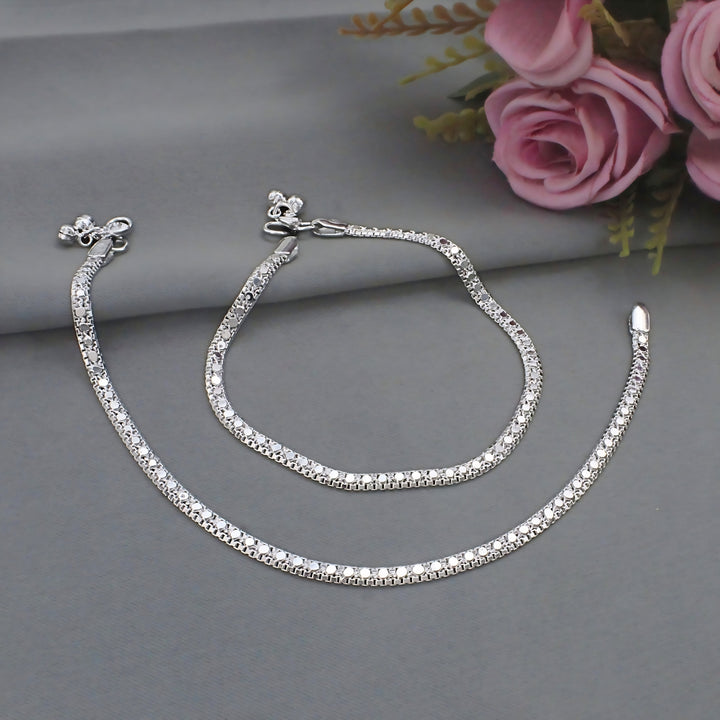 A pair of silver anklets with small, sparkling circular details is laid out on a smooth gray surface next to pink artificial roses.