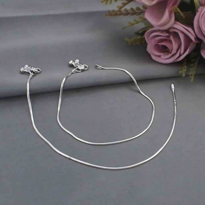 Elegant pair of silver snake chain anklets with delicate charms and tiny bells, beautifully displayed beside soft pink roses.