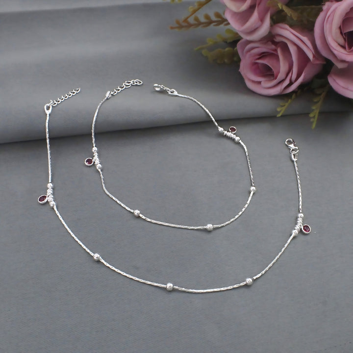 A pair of delicate silver anklets with tiny silver beads and small, round pink stone charms, displayed on a smooth grey surface next to pink roses.