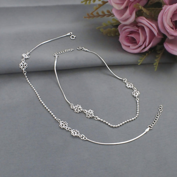 An image featuring two silver anklets, or anklet-style bracelets, gracefully laid out on a gray surface with pink roses in the upper right background.