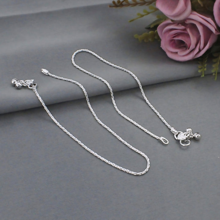 Two delicate silver anklets are laid out on a smooth gray surface next to a pink rose.