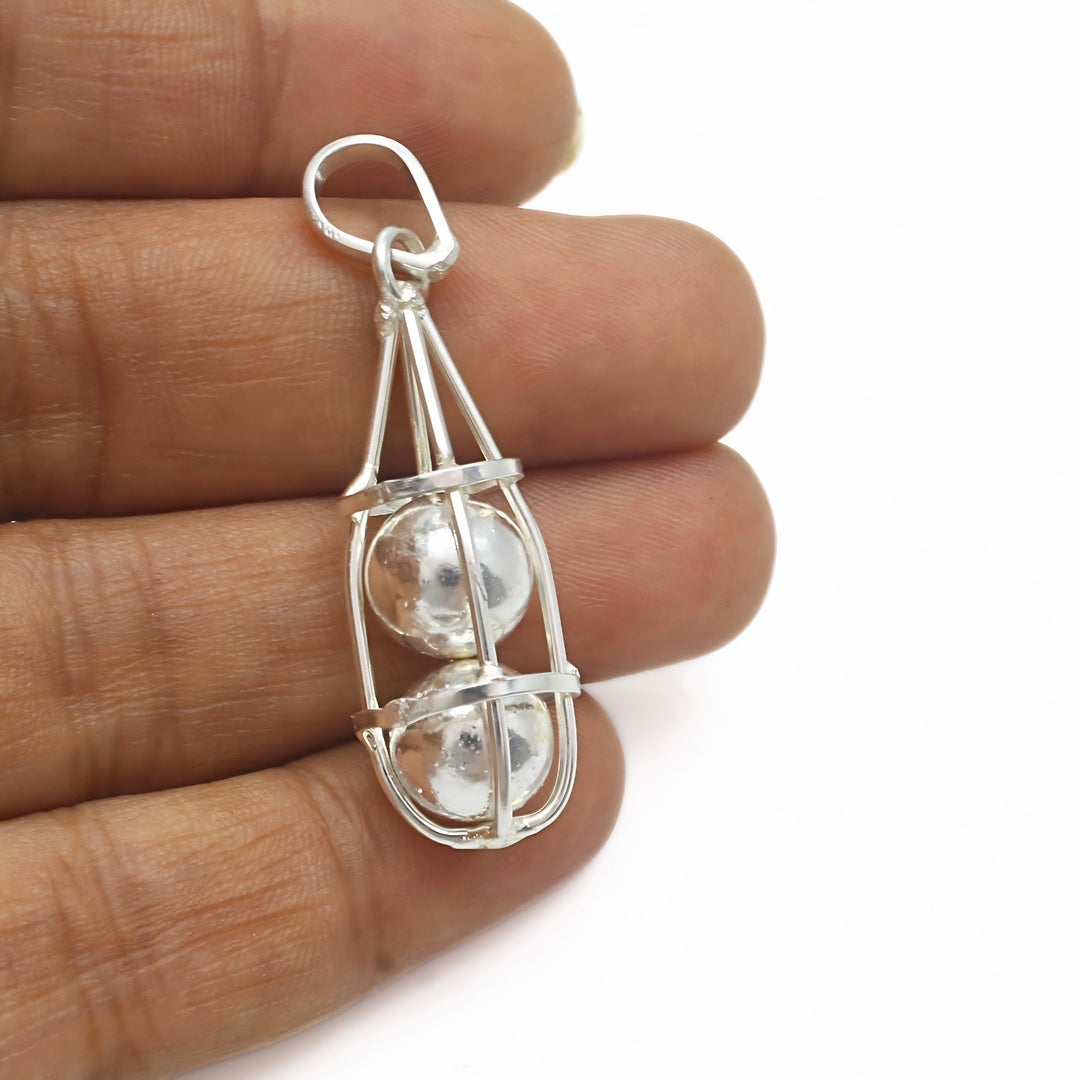 A close-up shot shows a silver pendant shaped like a cage, holding two silver balls inside, held between the fingers of a hand.