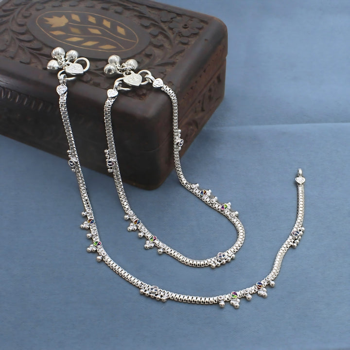A pair of silver anklets with tiny charms and small bells are draped over a dark wooden box on a blue surface.