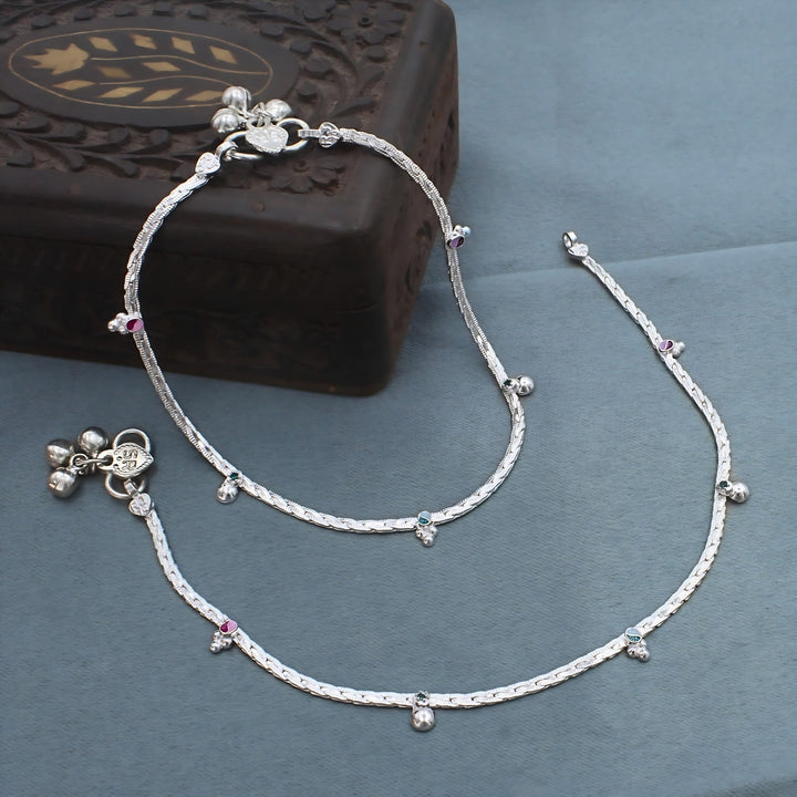 A pair of shiny silver anklets, also known as anklet, rests on a smooth grey surface next to a dark, carved wooden box.