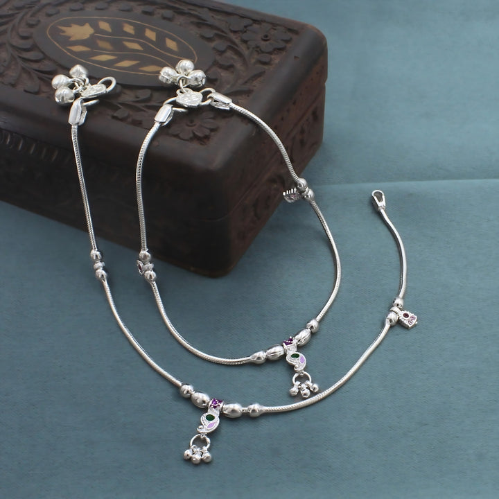 An image shows a pair of silver anklets with tiny charms and small hanging bells, neatly arranged on a blue surface next to a carved wooden box.