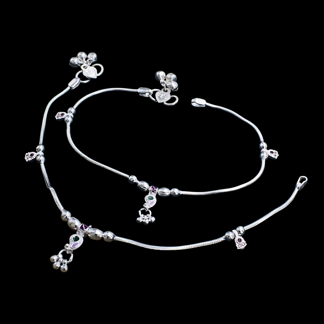 A pair of silver anklets with small charms and bells on a black background.
