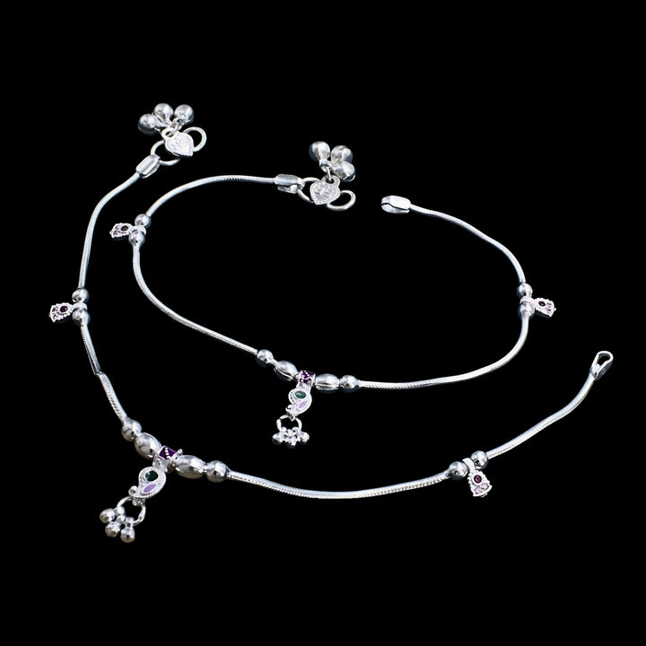 A pair of silver anklets with small charms and bells on a black background.