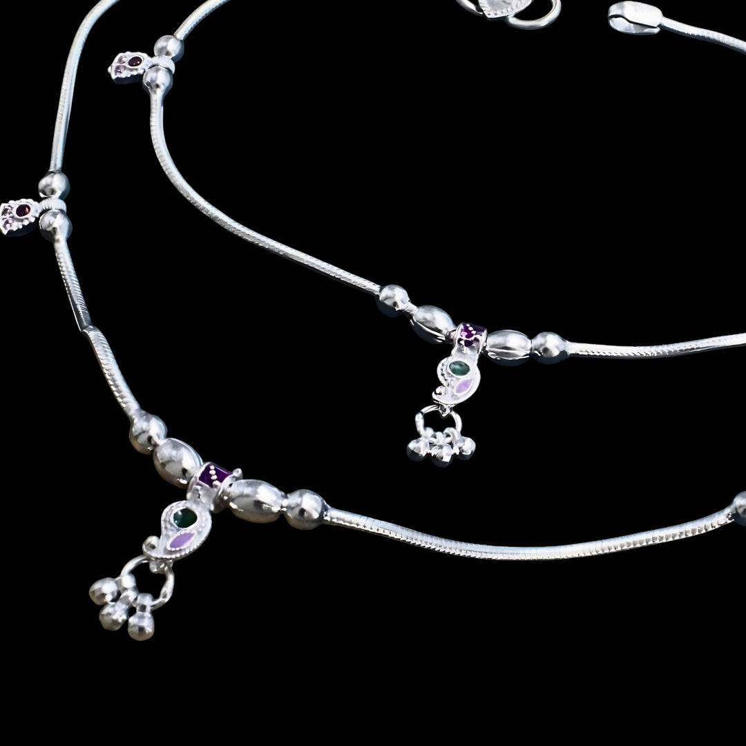 A pair of silver anklets with small charms containing green and purple enamel details, set against a black background.