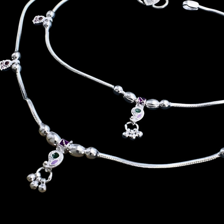 A pair of silver anklets with small charms containing green and purple enamel details, set against a black background.