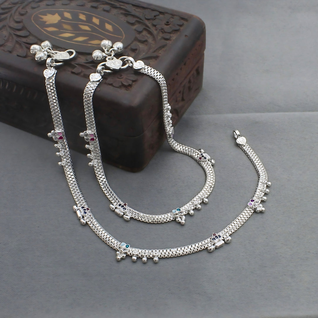 A pair of silver anklets with small colored stones and tiny bells is draped over and around a dark wooden box.