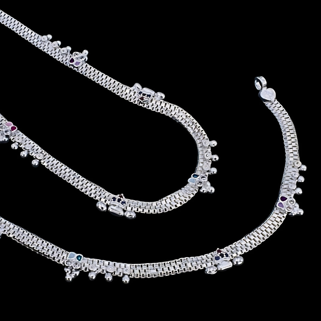 An image of silver anklets with tiny charms and small colored stones, set against a solid black background.