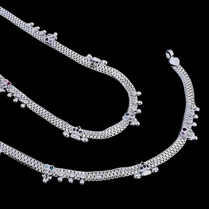 An image of silver anklets with tiny charms and small colored stones, set against a solid black background.