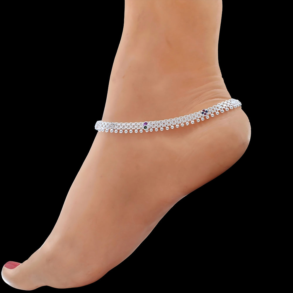 A person's foot and ankle are shown against a solid black background, featuring a silver anklet with intricate detailing and small colored stones.