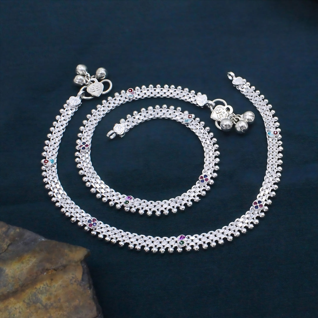 Intricate silver anklets with bead detailing, colorful stone accents, and ghungroo bells arranged in a spiral on a dark fabric background.