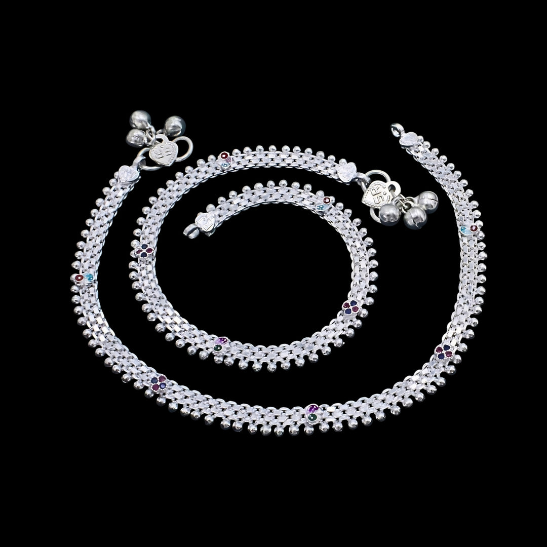 A pair of silver anklets with tiny bells and colorful accents, arranged in a spiral pattern against a solid black background.