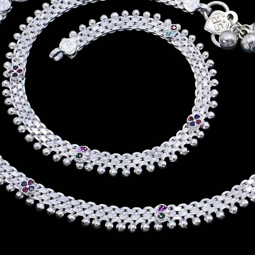 A high-angle, close-up shot shows a pair of silver anklets against a solid black background.
