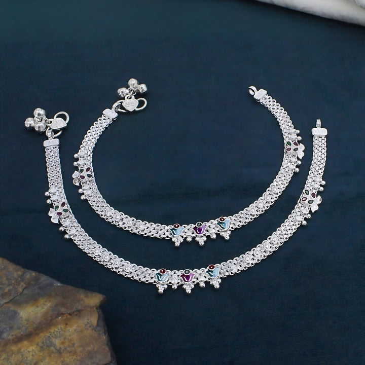 A pair of shiny silver Indian anklets, known as payal, are laid out in parallel curves against a dark teal background.