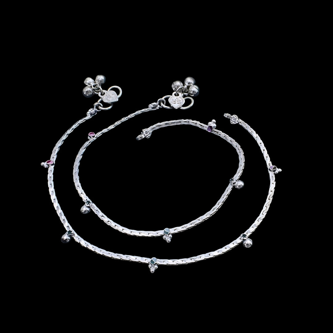 A pair of silver anklets with tiny charms and bells, displayed against a solid black background.