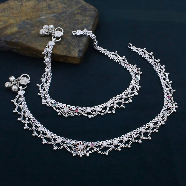 An overview of the image shows a pair of detailed silver anklets, known as payal, laid out against a dark, textured background.