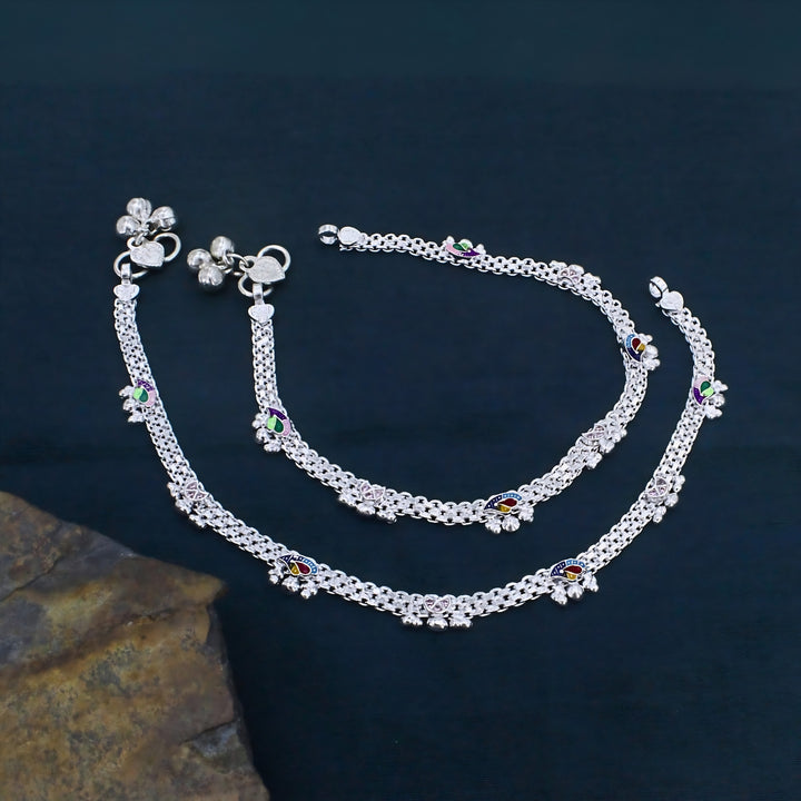 An overhead shot of a pair of traditional Indian silver anklets, known as payal, laid out on a dark blue surface next to a textured brown rock.
