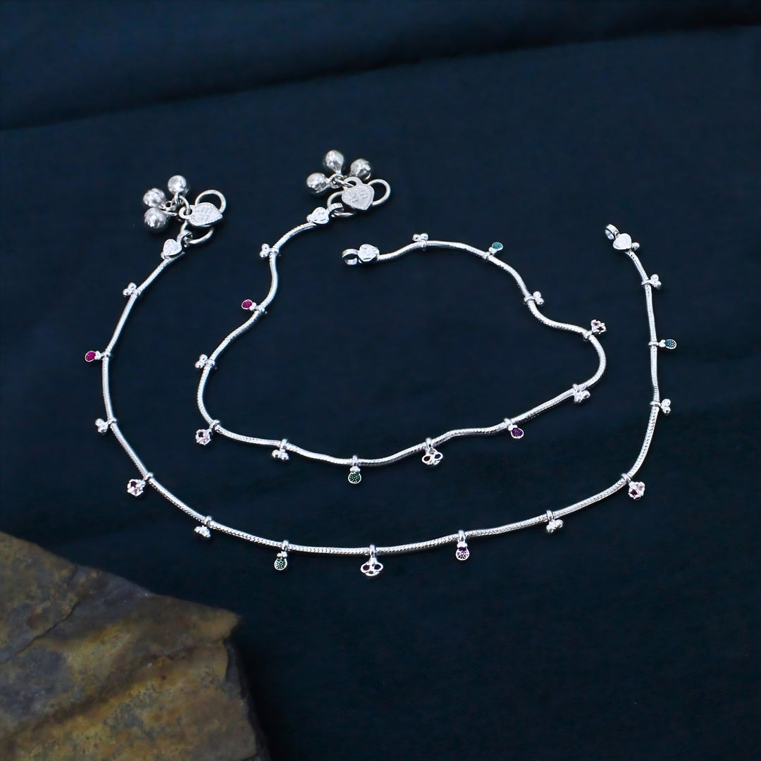 Two delicate silver anklets with tiny charms and small colored beads are neatly arranged on a smooth, dark blue background.