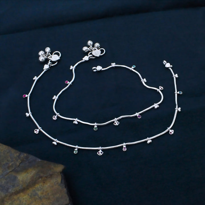 Two delicate silver anklets with tiny charms and small colored beads are neatly arranged on a smooth, dark blue background.