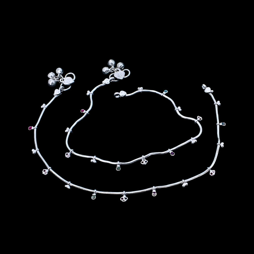 A pair of silver anklets with tiny charms and small bells, arranged in loops against a solid black background.