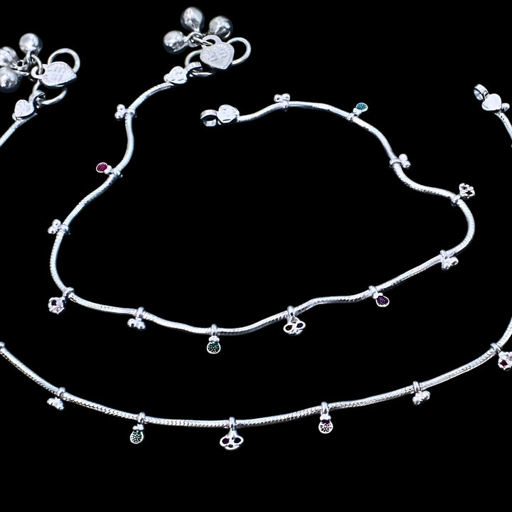 An image of a pair of silver anklets (payal) with small charms and colored stones, laid out against a black background.