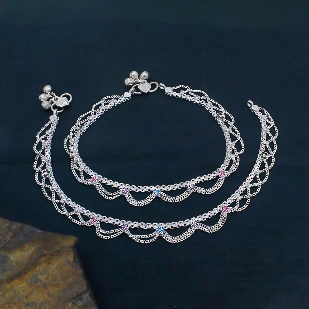 Layered silver anklets with colorful leaf accents and ghungroo bells displayed on a dark textured surface.