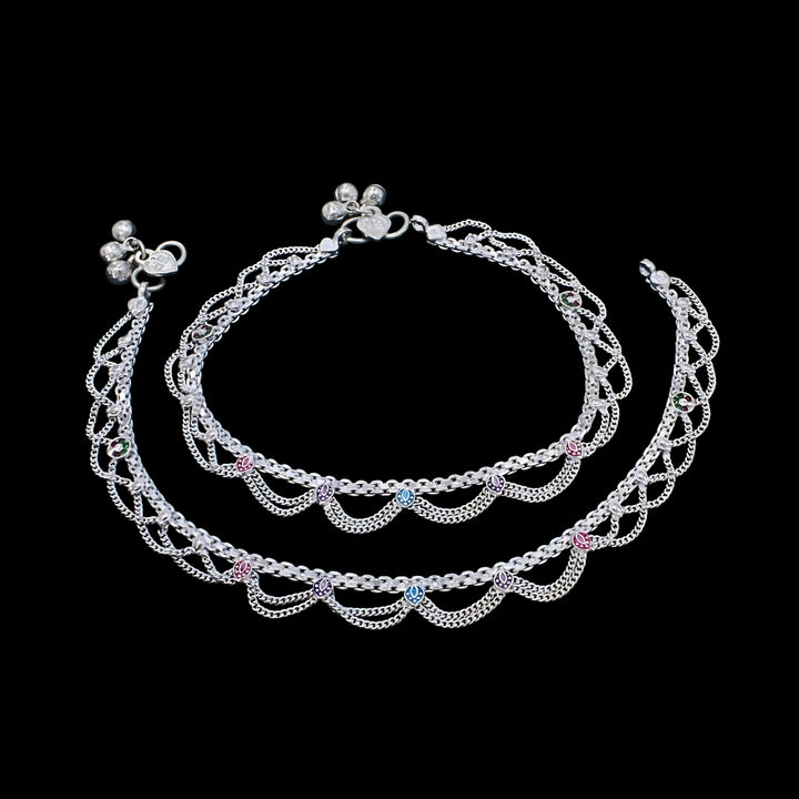 A pair of traditional silver anklets with delicate chain loops and small colorful charms, displayed against a solid black background.