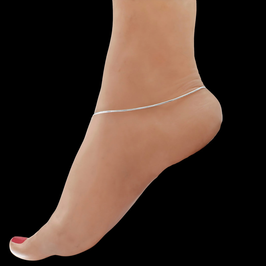 A side view of a person's bare foot against a black background, wearing a thin, shiny silver chain anklet.