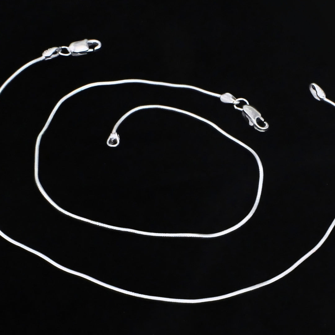 A bright silver snake chain anklet with a smooth, polished finish and secure lobster claw clasps, elegantly showcased on a black background.