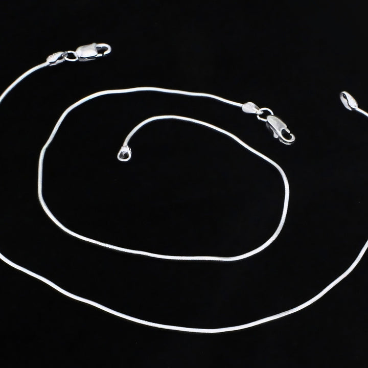 A bright silver snake chain anklet with a smooth, polished finish and secure lobster claw clasps, elegantly showcased on a black background.