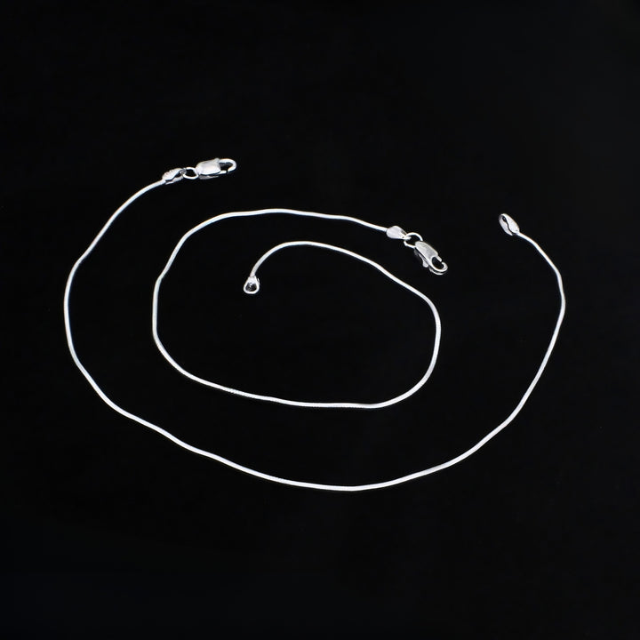 A silver snake chain anklet coiled on a solid black background, showing its smooth, round links and lobster claw clasps.