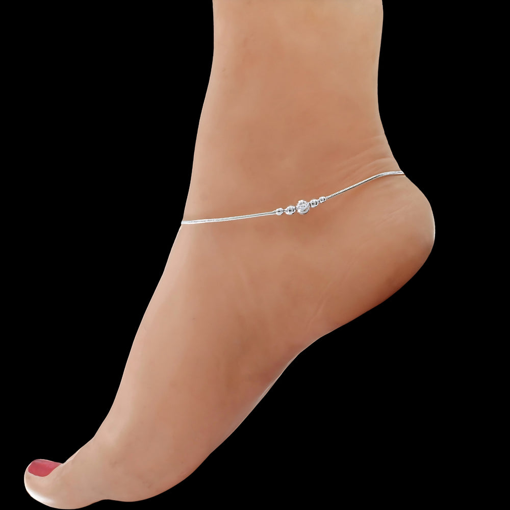 A close-up shot of a person's foot against a solid black background, showcasing a delicate silver anklet adorned with small beads and a central decorative charm.