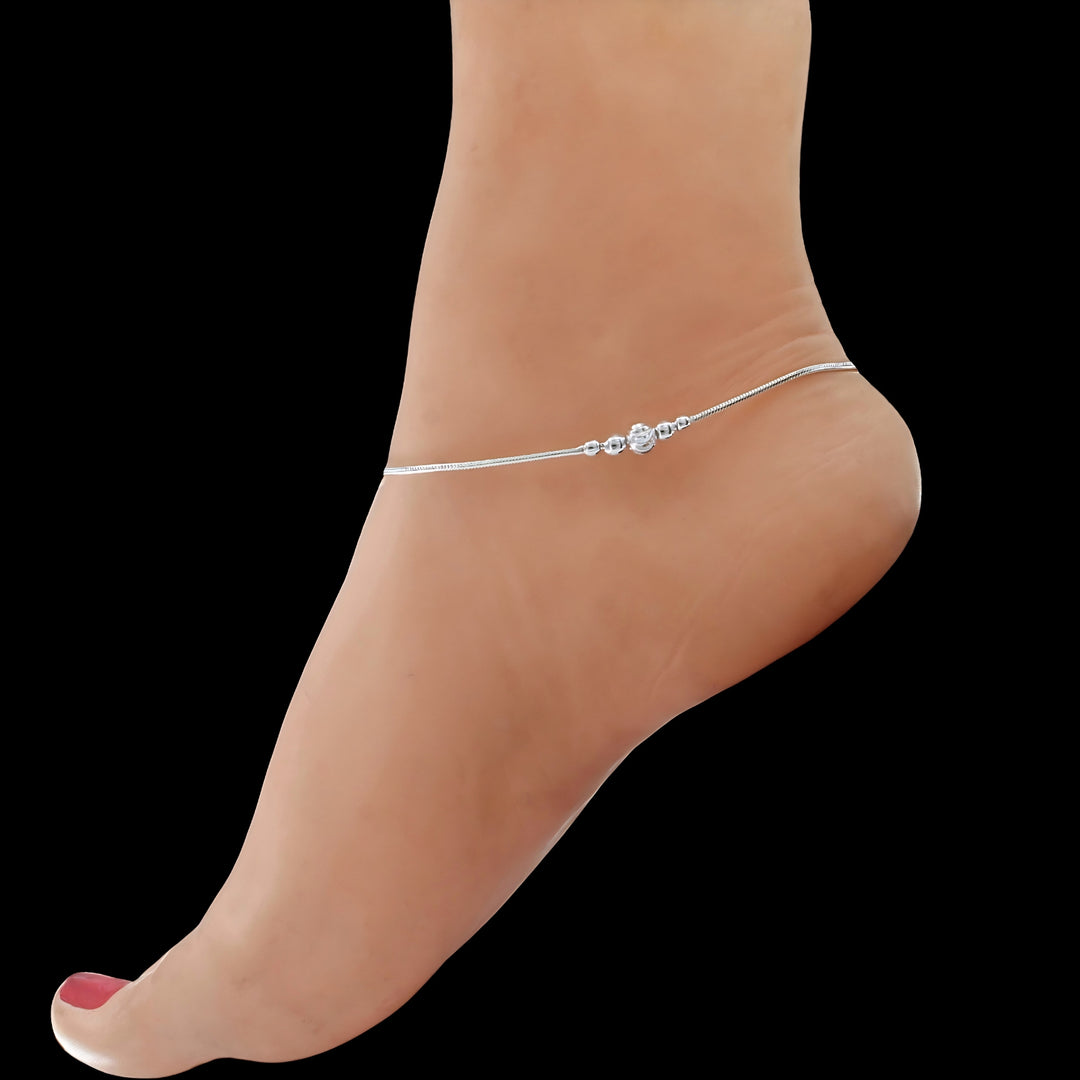 A close-up shot of a person's foot against a solid black background, showcasing a delicate silver anklet adorned with small beads and a central decorative charm.