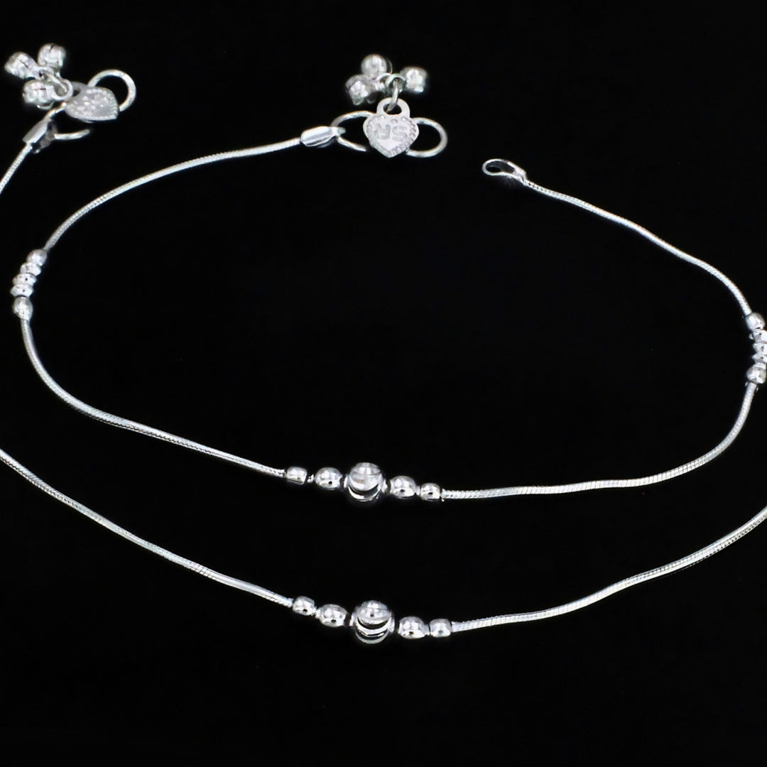 A pair of elegant silver anklets with smooth snake chains, delicate bead accents, central charms, and heart-and-flower detailed clasps, beautifully displayed on a black background.