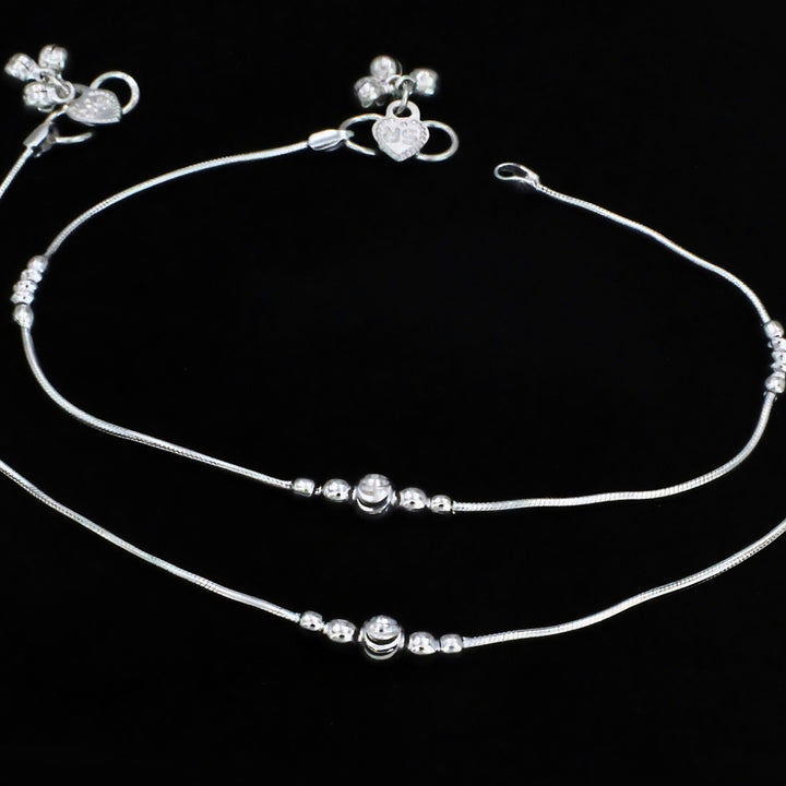 A pair of elegant silver anklets with smooth snake chains, delicate bead accents, central charms, and heart-and-flower detailed clasps, beautifully displayed on a black background.