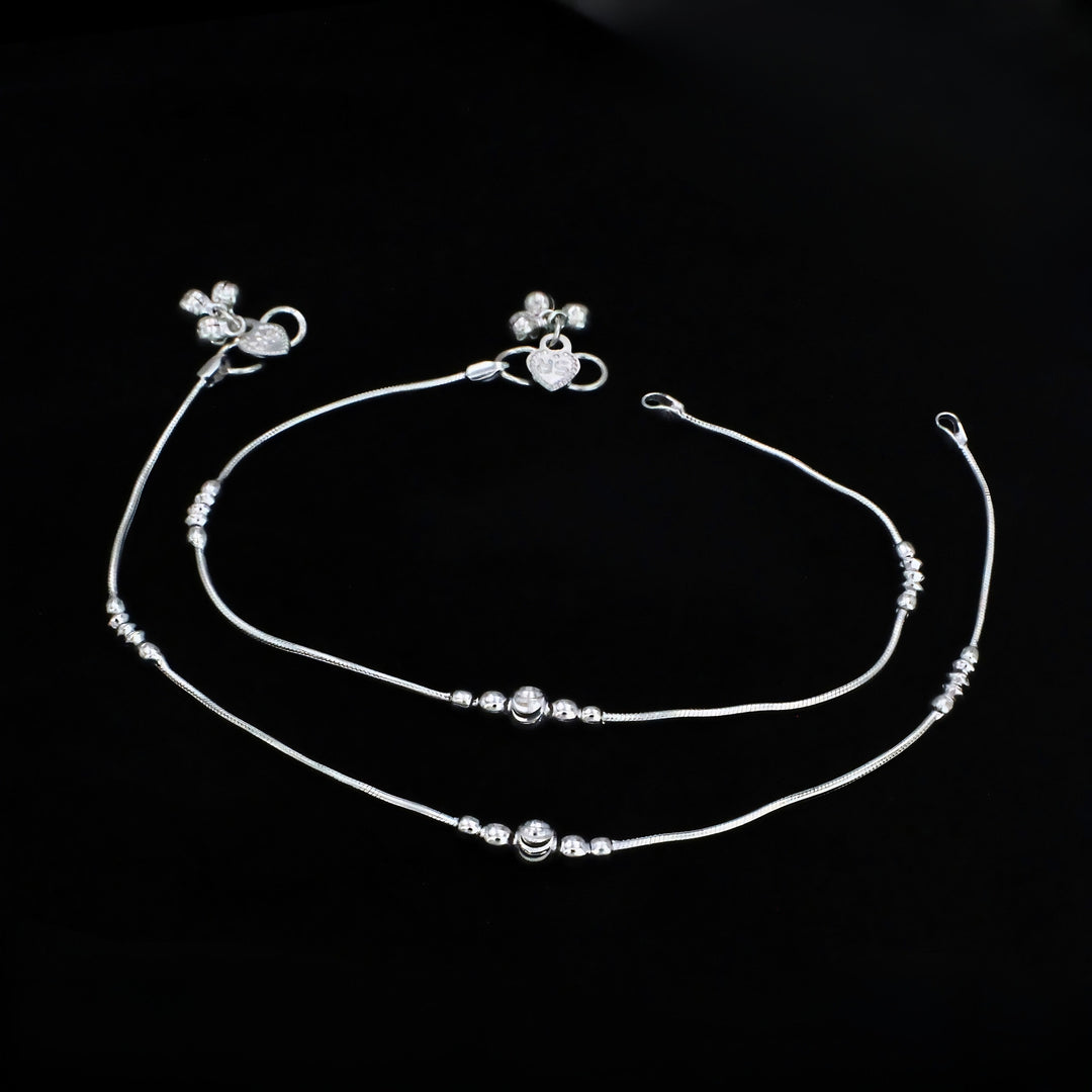 A pair of thin silver anklets with small decorative beads and charms, laid out horizontally against a solid black background.