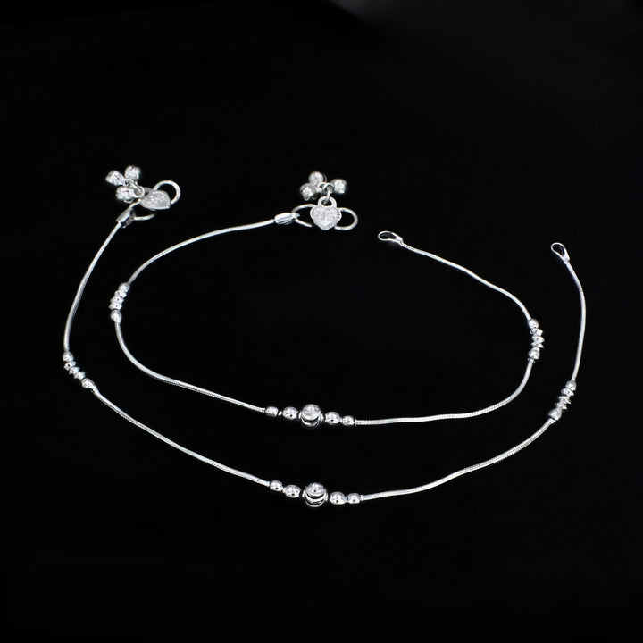 A pair of thin silver anklets with small decorative beads and charms, laid out horizontally against a solid black background.