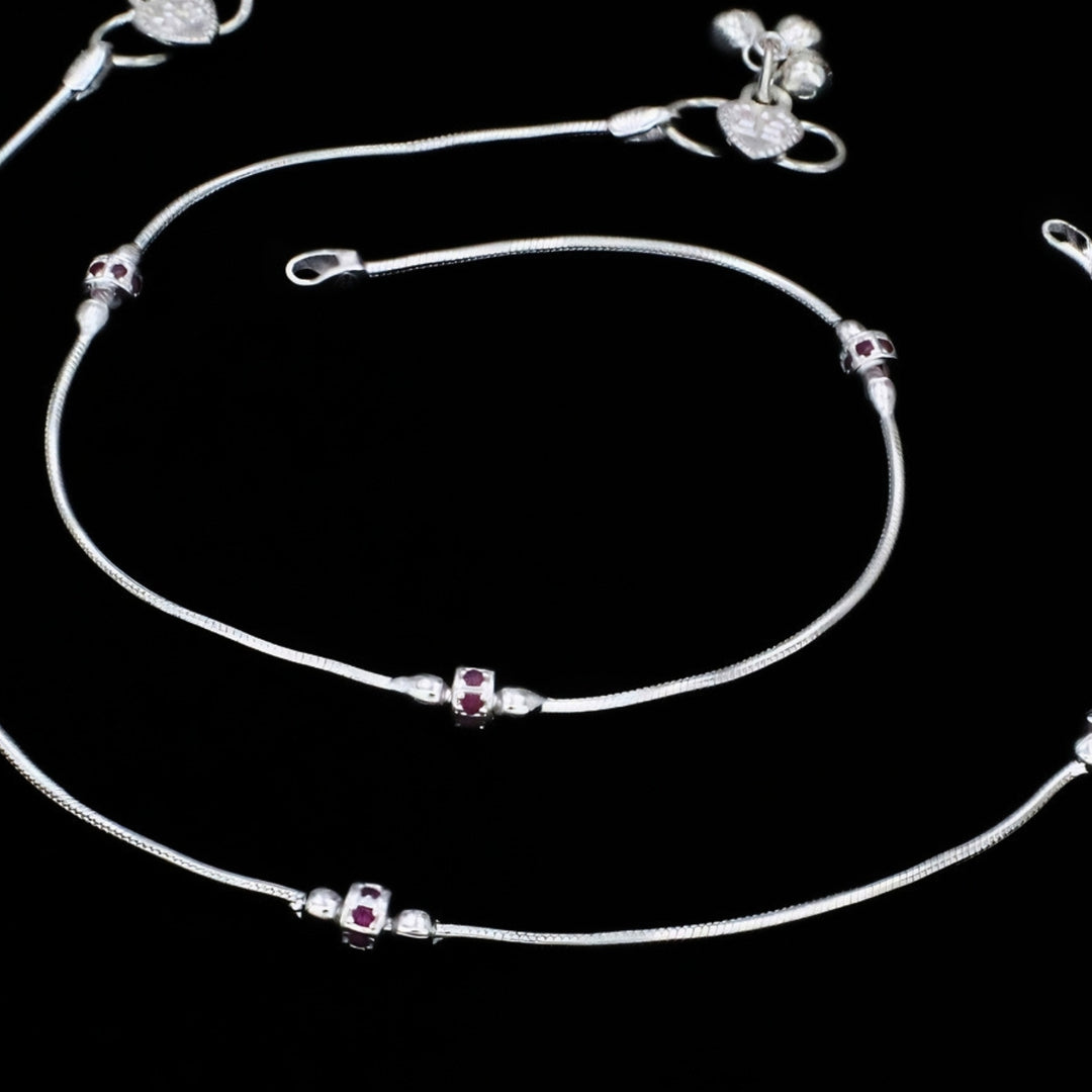A pair of silver snake chain anklets featuring small, decorative cylindrical charms inset with dark pink stones, set against a solid black background.