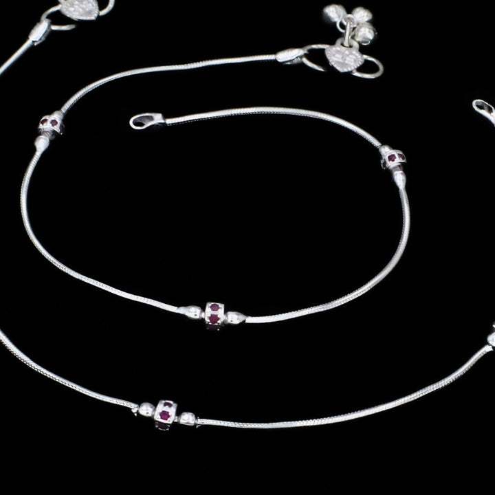 A pair of silver snake chain anklets featuring small, decorative cylindrical charms inset with dark pink stones, set against a solid black background.
