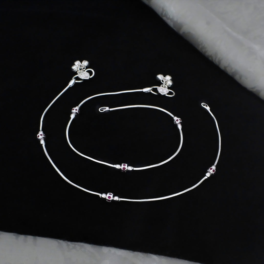 A pair of silver anklets with tiny charms rests on a black fabric surface.