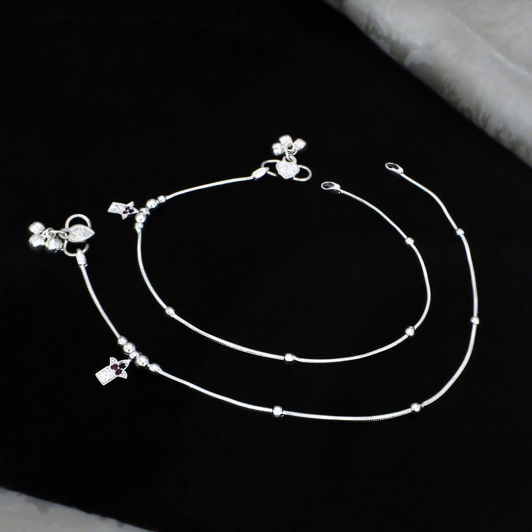 An elegant pair of thin silver anklets, also known as payal, is neatly arranged and displayed against a solid, deep black background.