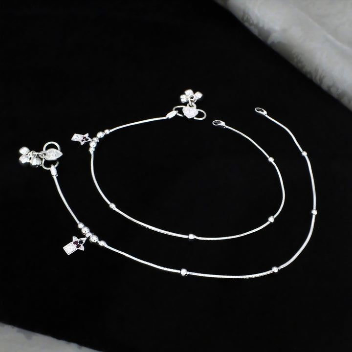 An elegant pair of thin silver anklets, also known as payal, is neatly arranged and displayed against a solid, deep black background.