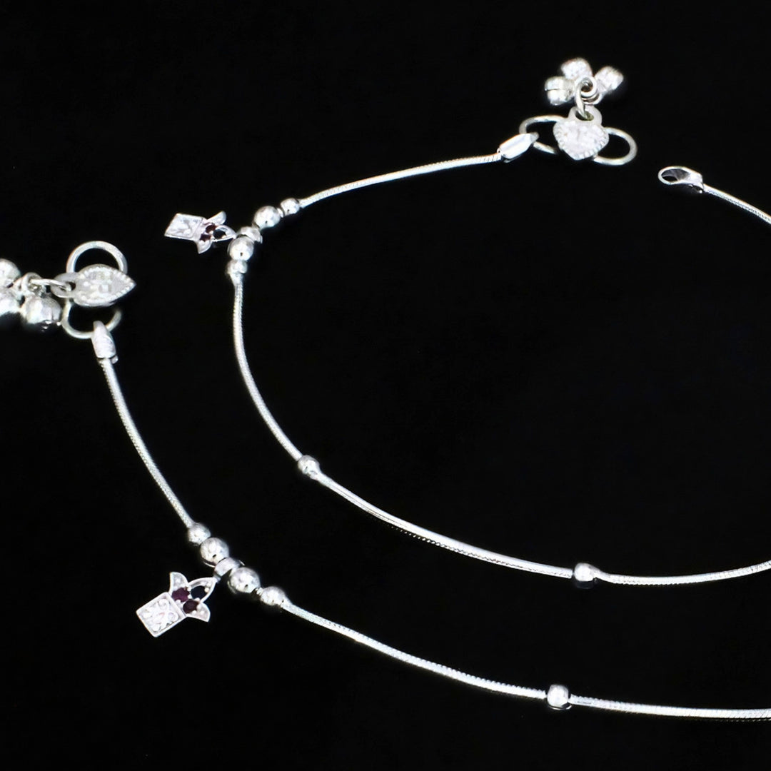 A pair of silver anklets with small charms and beads on a plain black background.