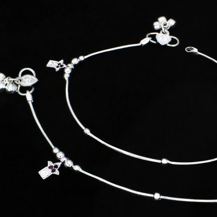 A pair of silver anklets with small charms and beads on a plain black background.