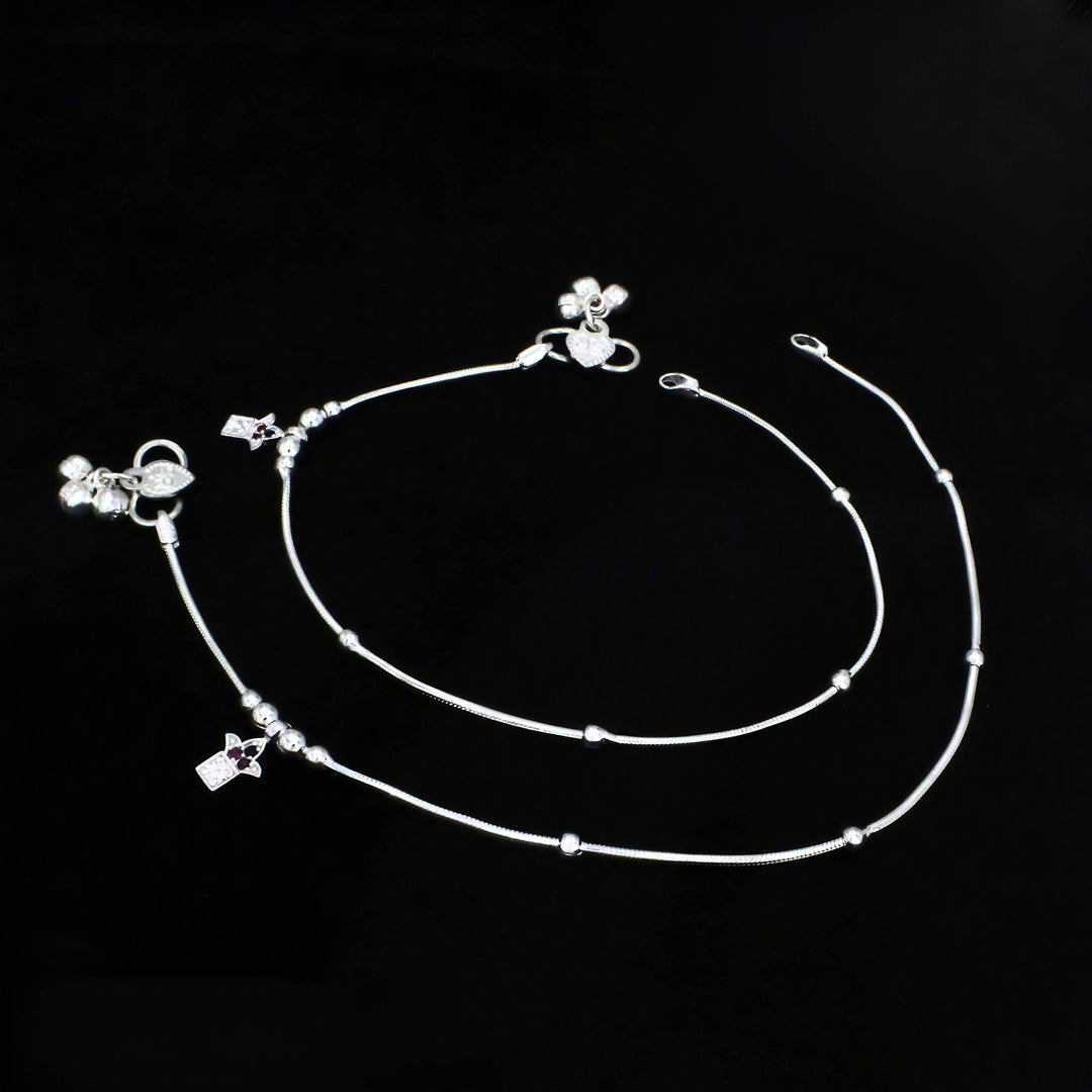 An overhead shot of a pair of delicate silver anklets laid flat against a solid black background.