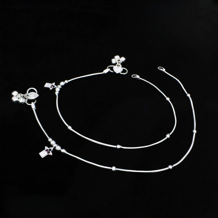 An overhead shot of a pair of delicate silver anklets laid flat against a solid black background.