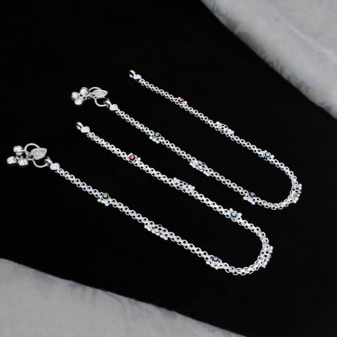 A close-up shot shows three silver anklets with tiny colored stones and small bells, neatly arranged on a black fabric surface.
